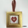 A gold framed cross stitch heart hanging on a red and green plaid ribbon.