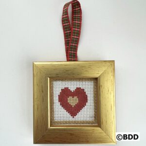 A gold framed cross stitch heart hanging on a red and green plaid ribbon.