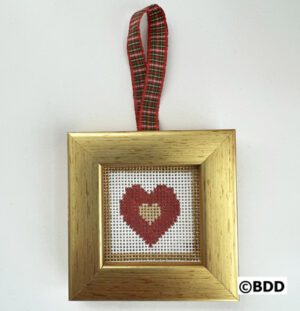 A gold framed cross stitch heart hanging on a red and green plaid ribbon.