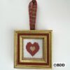 A cross stitch heart hanging in a gold frame.