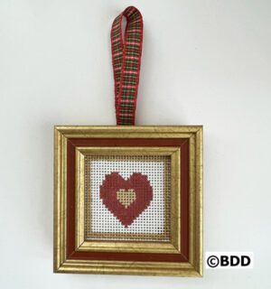 A cross stitch heart hanging in a gold frame.